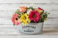 Colorful flower arrangement in a metal container labeled 'Flowers & Garden' on shiplap background.