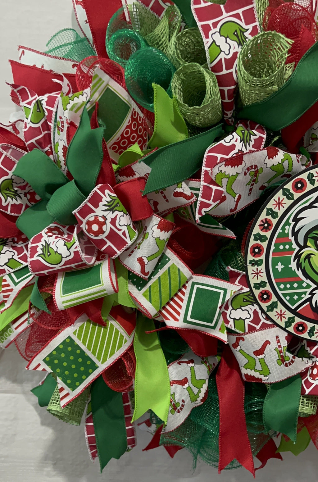 Handmade red and green Christmas wreath with a whimsical Grinch-inspired face sign, featuring festive ribbon and mesh for holiday front door decor