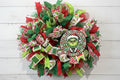 Handmade red and green Christmas wreath with a whimsical Grinch-inspired face sign, featuring festive ribbon and mesh for holiday front door decor.