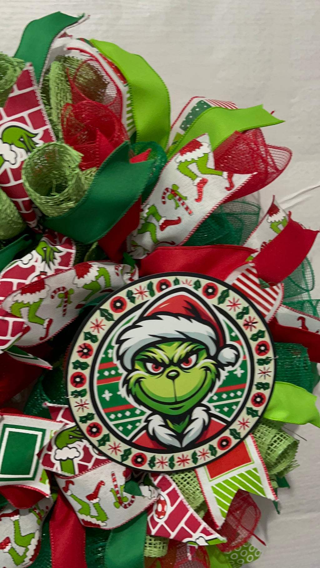 Handmade red and green Christmas wreath with a whimsical Grinch-inspired face sign, featuring festive ribbon and mesh for holiday front door decor
