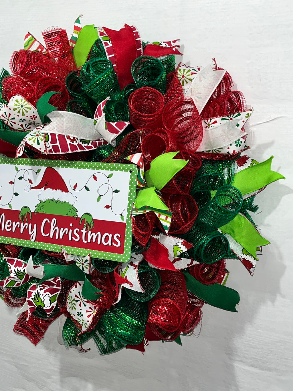 Handmade red and green Christmas wreath with a ‘Merry Christmas’ sign and whimsical Grinch-inspired design for festive front door decor.