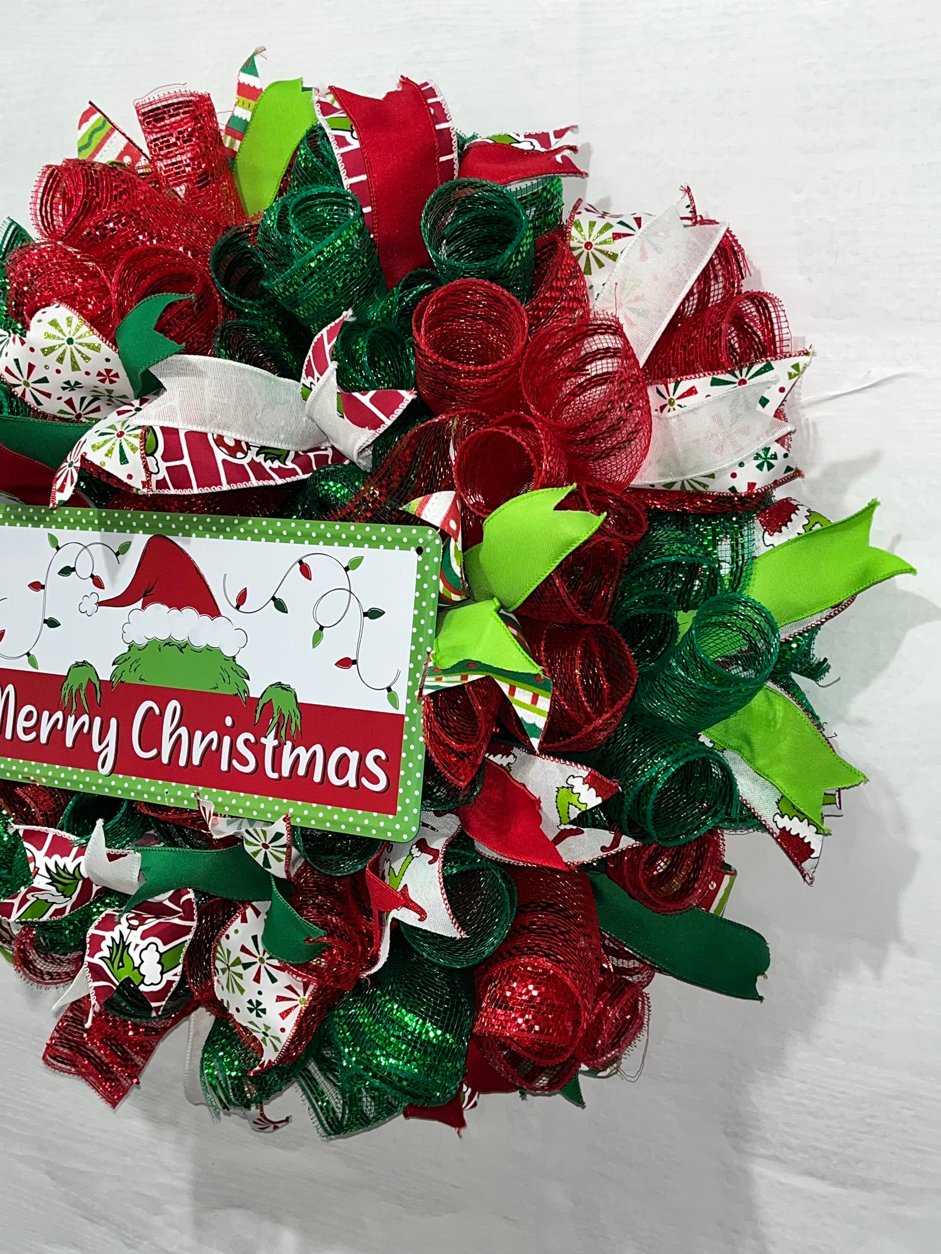 Handmade red and green Christmas wreath with a ‘Merry Christmas’ sign and whimsical Grinch-inspired design for festive front door decor.