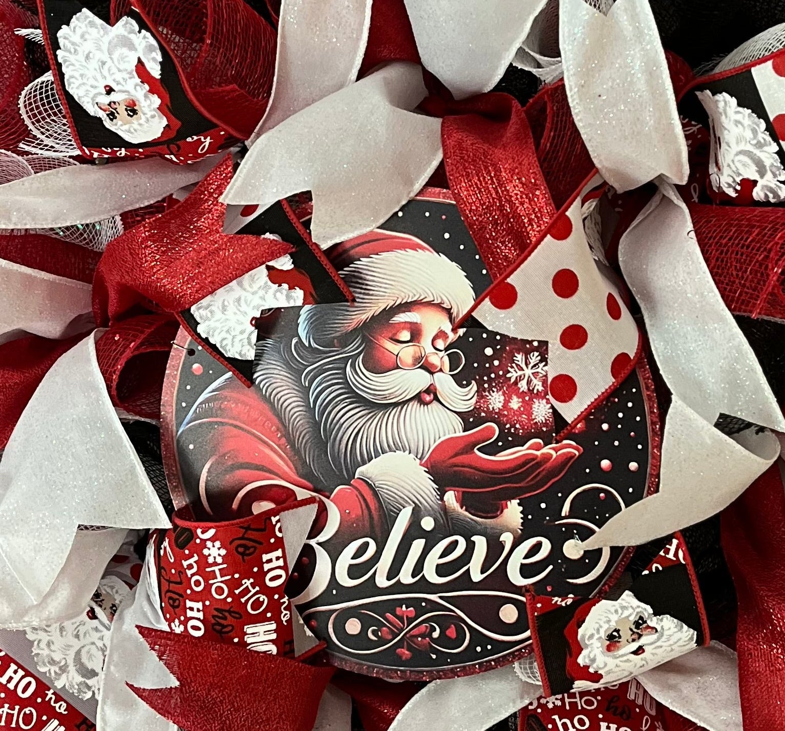 Handmade decorative Christmas wreath with red, white, and black ribbons and a Santa Claus "Believe" center sign.